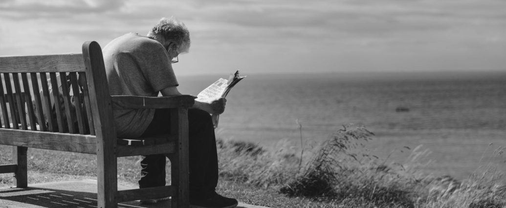 grayscale photo of man sitting on brown wooden bench reading news paper during day time