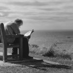 grayscale photo of man sitting on brown wooden bench reading news paper during day time